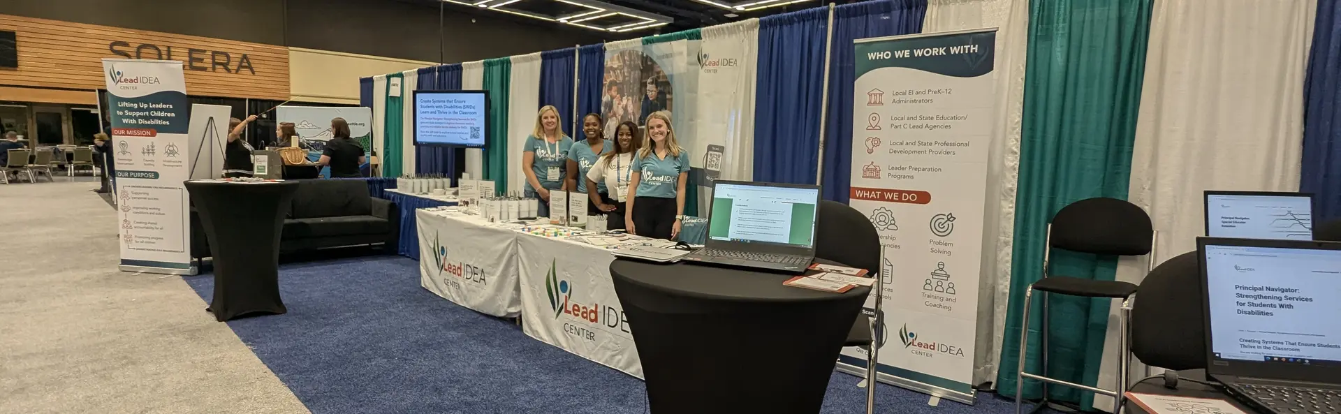Lead IDEA Center staff stand behind tables facing the camera and smiling at a conference.