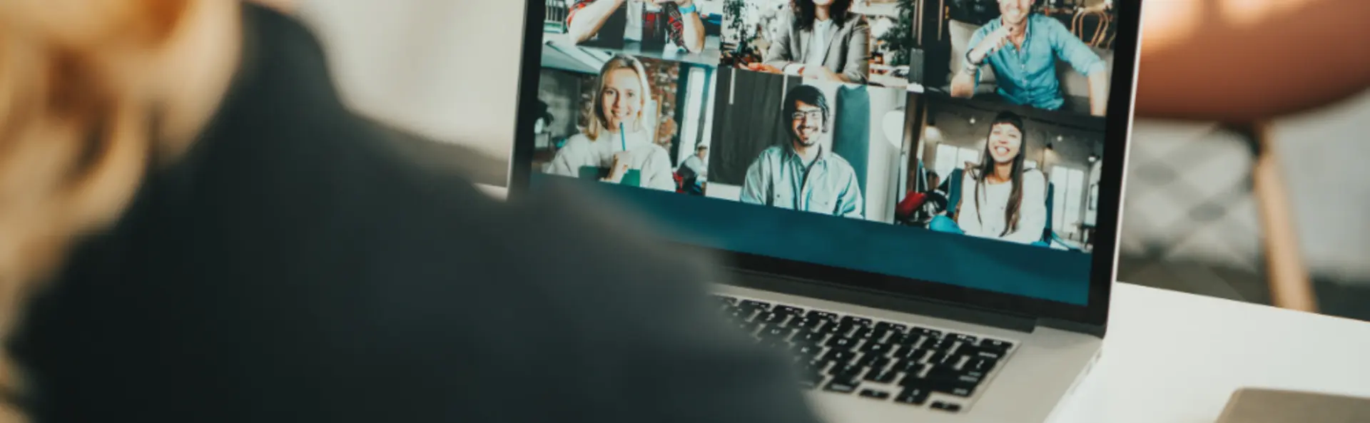 Person sits at table and looks at laptop screen which shows a video call with six people