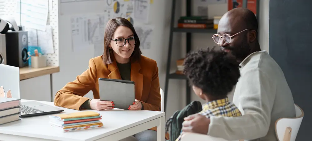 A student and a parent sit at a desk, facing towards an educational leader.